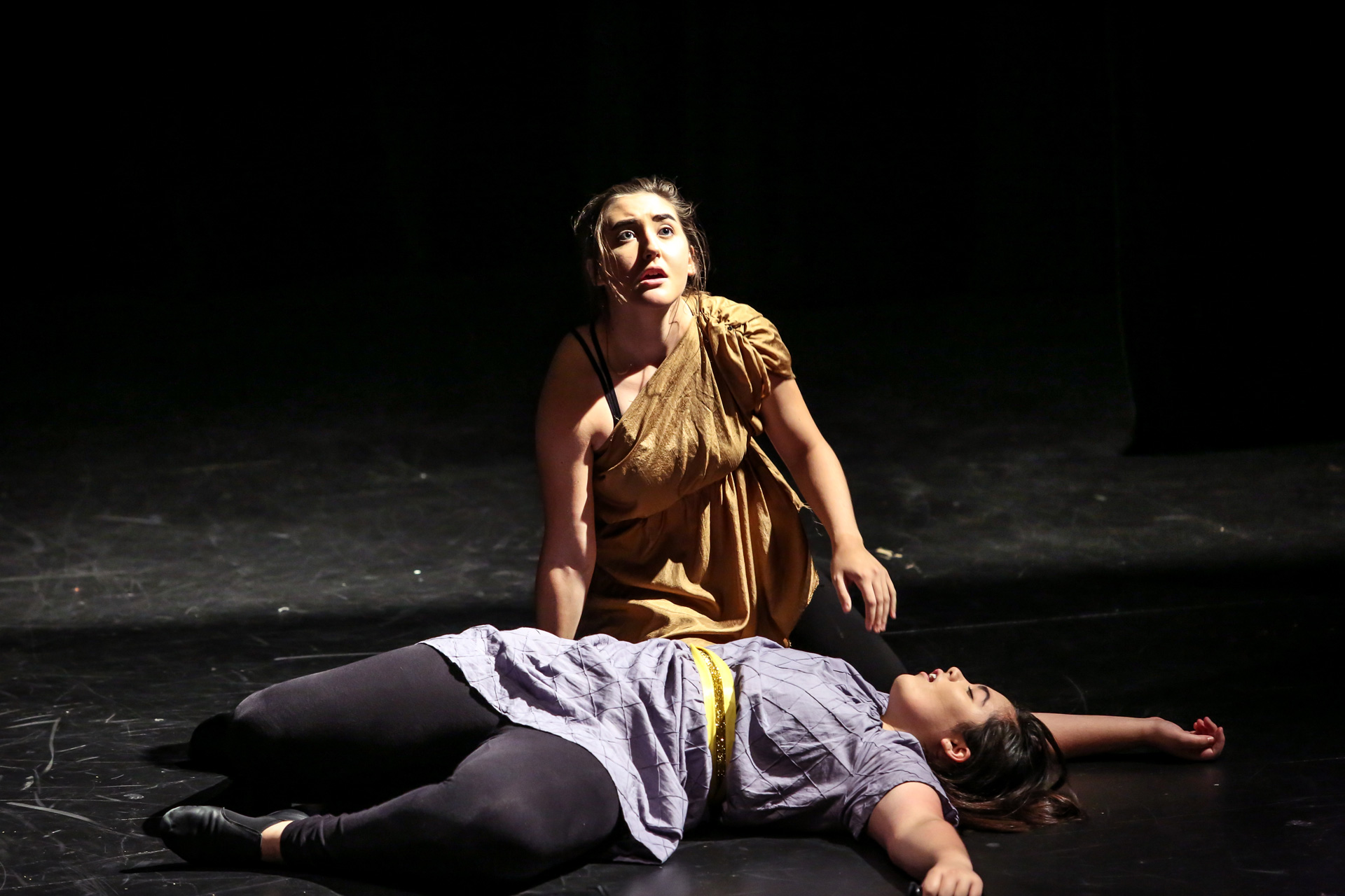 Patel students on stage, one kneeling on the ground looking up and the other laying on the ground.