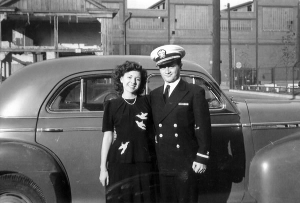 A black and white photo of a couple posing in front of a car.
