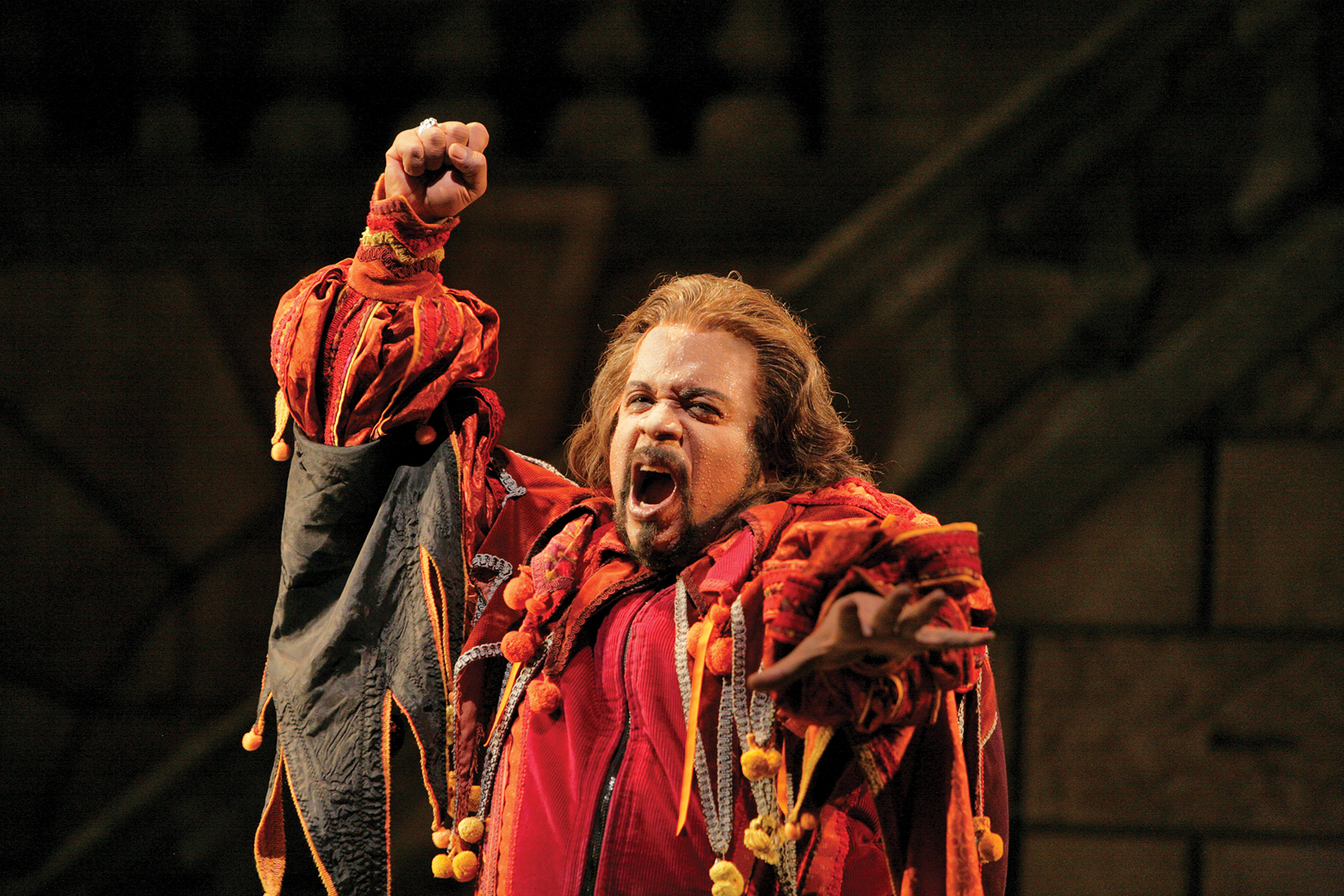 Opera performer in a red costume singing with fist raised in the air and arm outstretched to the audience.